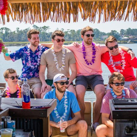 Group of seven friends wearing leis and sunglasses, laughing and holding drinks on a tiki-style pontoon boat by a coastal marsh on a sunny summer day.