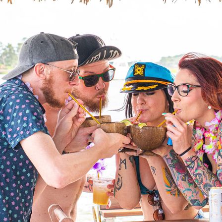 Four friends on a boat deck sipping coconut cocktails through straws, wearing leis, sunglasses and a captain's hat at a sunny tropical waterfront party