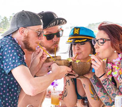 Four friends on a boat deck sipping coconut cocktails through straws, wearing leis, sunglasses and a captain's hat at a sunny tropical waterfront party