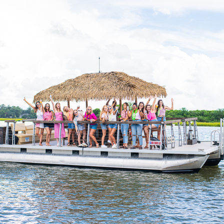 Group of friends celebrating on a tiki-roof pontoon boat cruising a coastal waterway, wearing summer outfits and leis