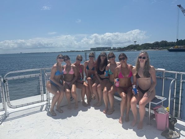 Nine friends in colorful bikinis sitting on a boat deck holding drinks, smiling toward the camera with a sunny coastal bay and shoreline skyline in the background.