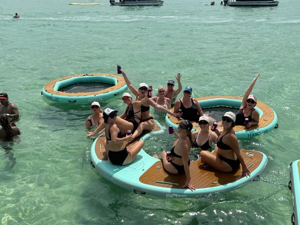Group of women in bikinis laughing and holding drinks on round inflatable floating platforms in clear turquoise shallow ocean, with boats in the distance for a sunny beach-party vibe.