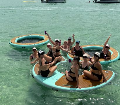 Group of women in bikinis laughing and holding drinks on round inflatable floating platforms in clear turquoise shallow ocean, with boats in the distance for a sunny beach-party vibe.