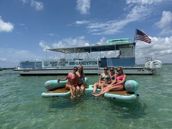 Five friends in bright bikinis lounging on inflatable boards in clear turquoise shallows beside a tour boat flying an American flag under a sunny sky.