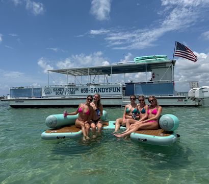 Five friends in bright bikinis lounging on inflatable boards in clear turquoise shallows beside a tour boat flying an American flag under a sunny sky.