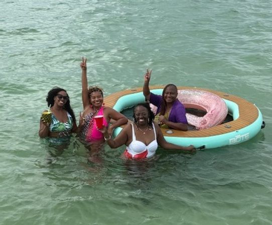 Four friends in colorful swimsuits smiling and holding drinks while standing in shallow green water next to a round aqua inflatable float with a pink donut tube; two flash peace signs for a fun summer beach float moment.