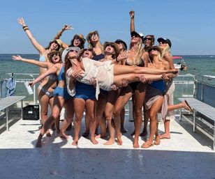 Group of women in swimsuits and sun hats laughing on a pontoon boat over blue water, lifting a friend in a joyful summer boat party on a sunny day