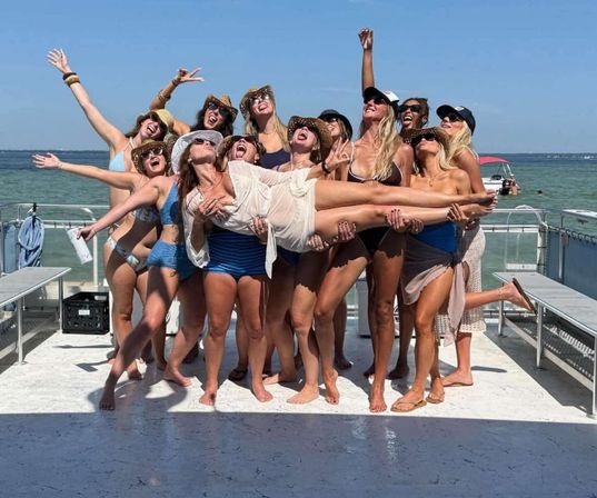 Group of women in swimsuits and sun hats laughing on a pontoon boat over blue water, lifting a friend in a joyful summer boat party on a sunny day