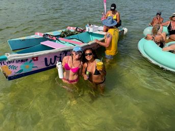 Two women in bikinis wading in shallow green coastal water holding tropical drinks beside a brightly painted small boat with other people on inflatable tubes nearby — summer beach party scene