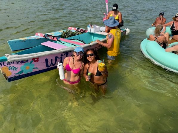 Two women in bikinis wading in shallow green coastal water holding tropical drinks beside a brightly painted small boat with other people on inflatable tubes nearby — summer beach party scene