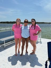 Three friends in sunglasses and colorful tees standing barefoot on a pontoon boat deck with calm bay water and a tree-lined shore under a sunny blue sky
