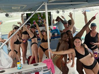 Lively boat party on a covered pontoon at the shoreline — group of adults in swimwear cheering and posing with drinks while a TV broadcasts a football game.