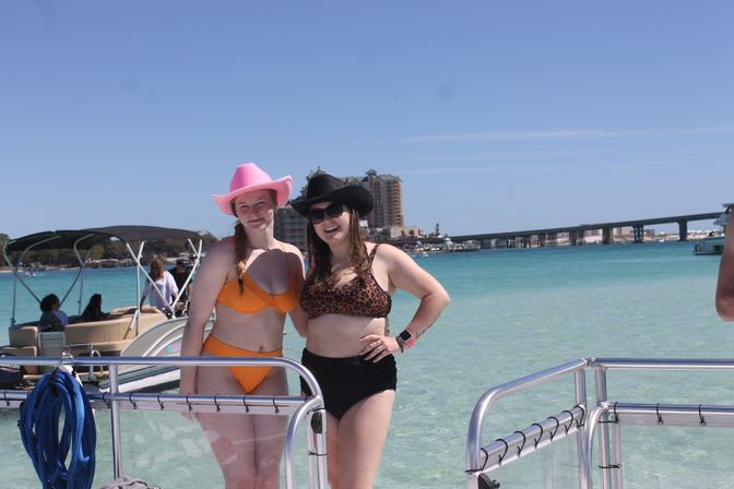 Two women in swimsuits and cowboy hats posing on a boat railing in clear turquoise beach water with a long pier and beachfront buildings under a sunny blue sky