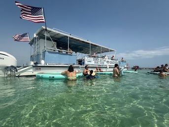 Tour boat with American flags anchored at a sunny sandbar, people on inflatables and wading in shallow clear turquoise water
