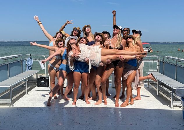 Group of women in swimsuits and sun hats holding a friend aloft on a sunny boat deck with turquoise coastal waters and clear blue sky