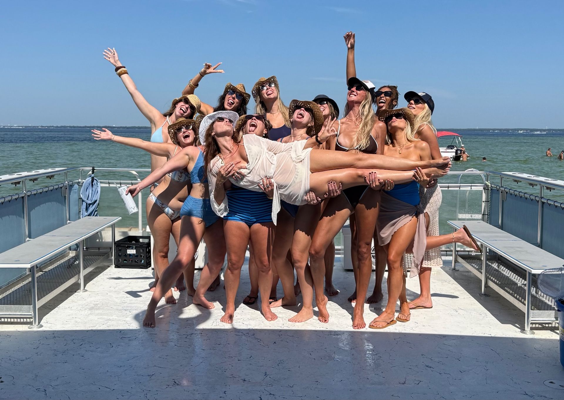 Group of women in swimsuits and sun hats holding a friend aloft on a sunny boat deck with turquoise coastal waters and clear blue sky