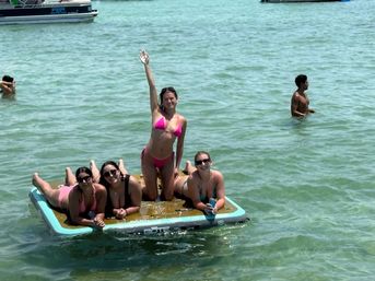 Four women in bikinis lounging and posing on an inflatable floating board in clear shallow green bay water with boats in the background on a sunny day