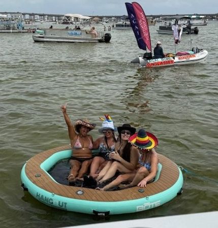 Four women in swimsuits and colorful hats lounging on a round inflatable floating island in a busy coastal bay with pontoon boats and advertising flags in the background — summer boating party