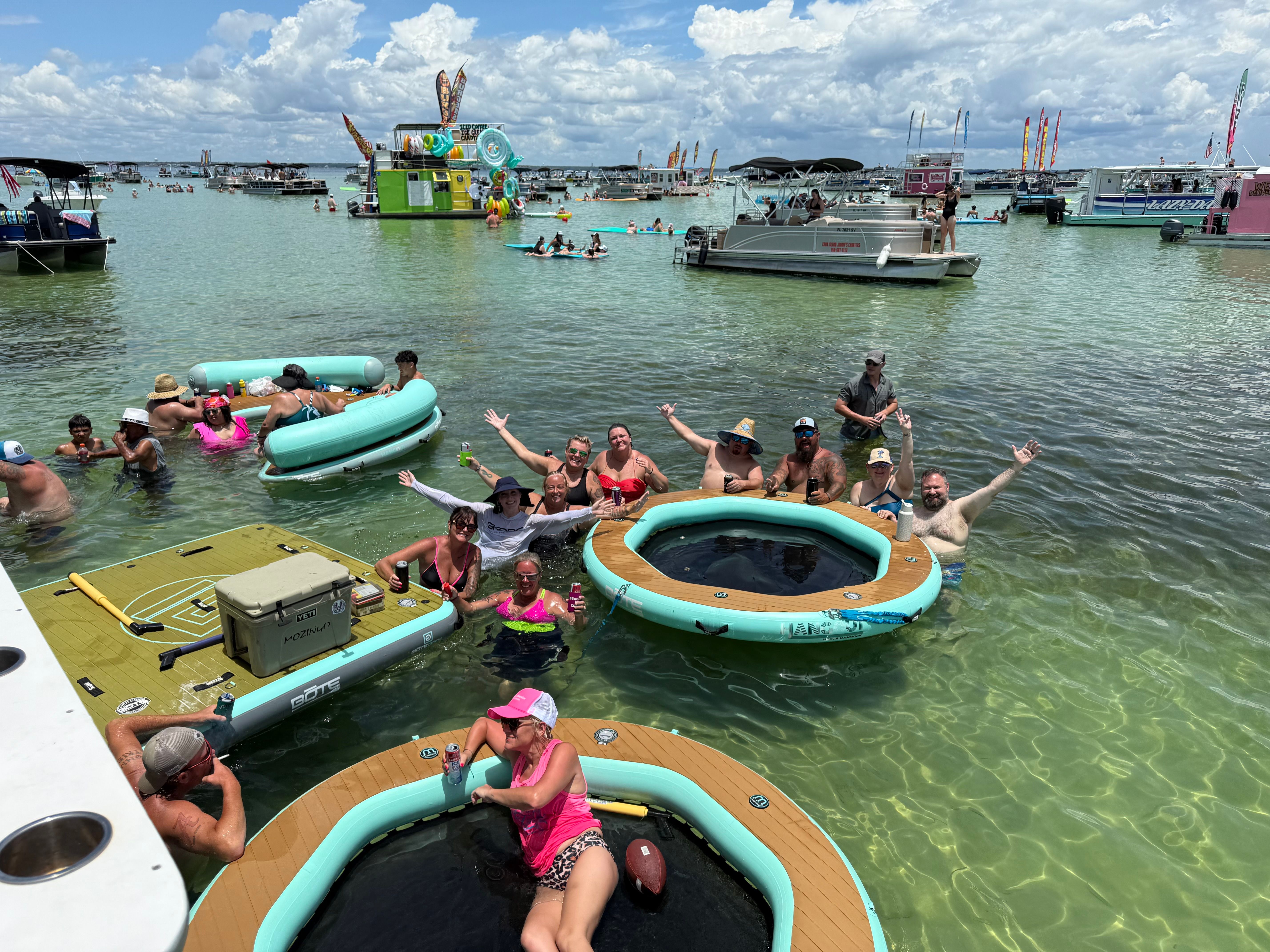 Shallow turquoise bay sandbar party with people relaxing on circular inflatable loungers, paddleboards and anchored pontoons, surrounded by party boats under a sunny sky with puffy clouds.