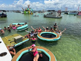 Shallow turquoise bay sandbar party with people relaxing on circular inflatable loungers, paddleboards and anchored pontoons, surrounded by party boats under a sunny sky with puffy clouds.