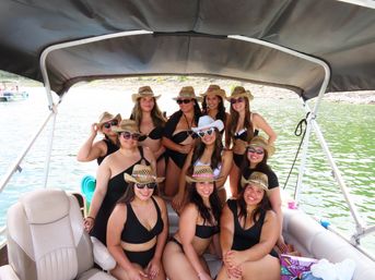 Group of women in black swimsuits and straw cowboy hats smiling under a canopy on a pontoon boat on a sunny green lake.