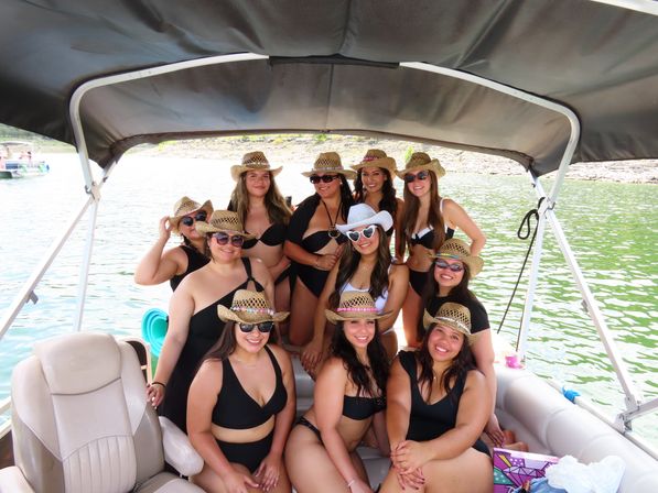 Group of women in black swimsuits and straw cowboy hats smiling under a canopy on a pontoon boat on a sunny green lake.