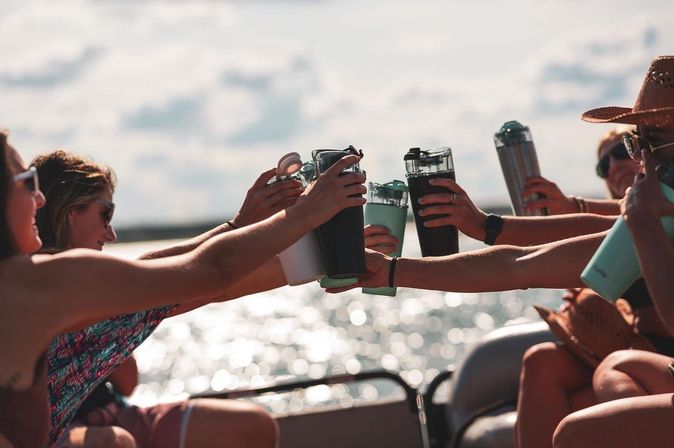 Friends on a sunny boat ride clinking insulated tumblers in a celebratory toast over sparkling lake water.