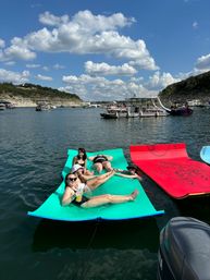 Group of friends lounging on bright floating mats at a busy summer lake, surrounded by boats, a multi‑level pontoon and rocky shoreline under a blue sky dotted with puffy clouds.