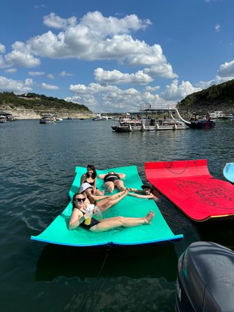 Group of friends lounging on bright floating mats at a busy summer lake, surrounded by boats, a multi‑level pontoon and rocky shoreline under a blue sky dotted with puffy clouds.