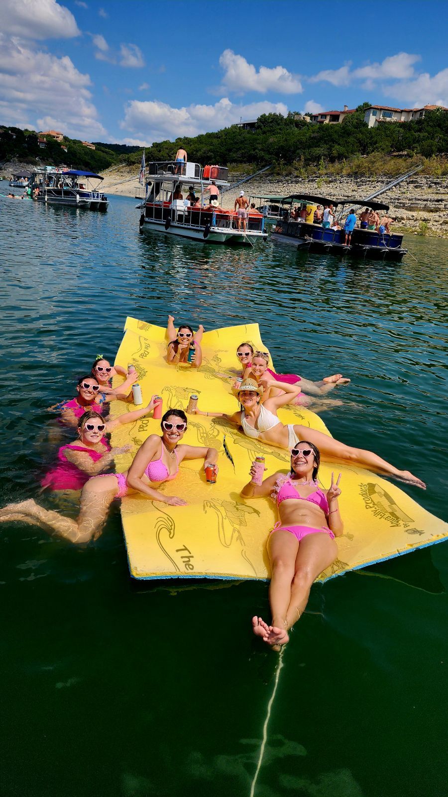 Group of friends in pink swimsuits relaxing and posing on a large yellow floating mat in a sunny lake, holding drinks and wearing heart-shaped sunglasses, with pontoon boats and a rocky tree-lined shoreline with summer homes in the background.