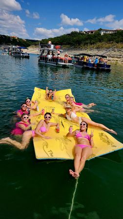 Group of friends in pink swimsuits relaxing and posing on a large yellow floating mat in a sunny lake, holding drinks and wearing heart-shaped sunglasses, with pontoon boats and a rocky tree-lined shoreline with summer homes in the background.
