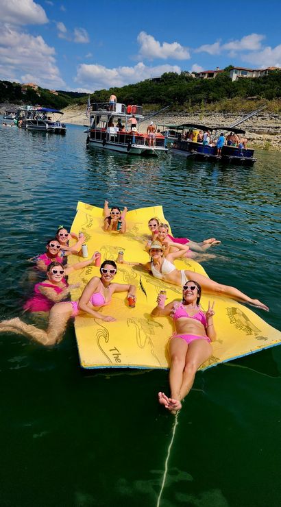 Group of friends in pink swimsuits relaxing and posing on a large yellow floating mat in a sunny lake, holding drinks and wearing heart-shaped sunglasses, with pontoon boats and a rocky tree-lined shoreline with summer homes in the background.