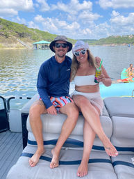 Happy couple on a pontoon boat enjoying a sunny lake day — both wearing sun hats and sunglasses; woman in a white bikini holding a canned drink with a straw, man in a navy long-sleeve shirt and striped swim shorts, green shoreline and docks in the background.