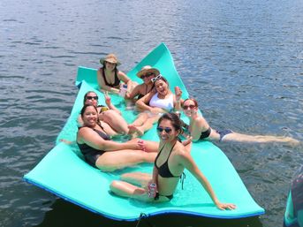 Seven friends in swimsuits lounging on a bright turquoise floating mat on a sunny lake, sipping drinks and smiling.