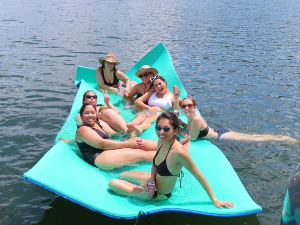 Seven friends in swimsuits lounging on a bright turquoise floating mat on a sunny lake, sipping drinks and smiling.