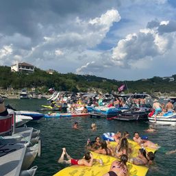 Crowded summer lake boat party with anchored motorboats, people swimming and lounging on a large yellow inflatable, paddleboards and jet skis near a wooded shoreline and homes under dramatic clouds.