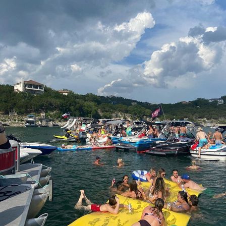 Crowded summer lake boat party with anchored motorboats, people swimming and lounging on a large yellow inflatable, paddleboards and jet skis near a wooded shoreline and homes under dramatic clouds.