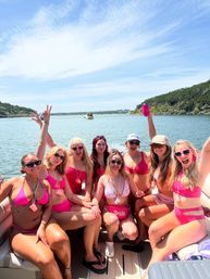 Cheerful group of women in pink swimwear on a pontoon boat at a sunny lake, celebrating a bachelorette with drinks and a woman wearing a 'Bride' sash