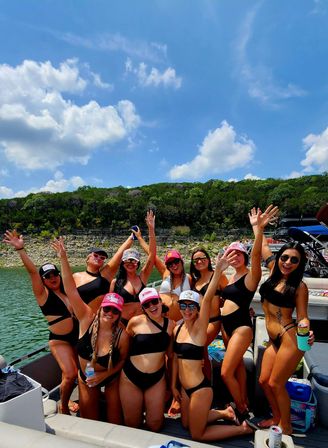 Group of friends in black swimsuits and colorful caps cheering on a sunlit party boat at a green, rocky lakeshore under a bright blue summer sky