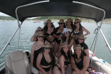 Group of friends in black swimsuits and straw cowboy hats smiling together on a covered pontoon boat at a sunny lake with wooded shoreline in the background.