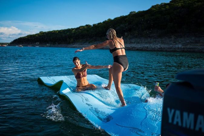 Two friends in swimsuits playing on a blue inflatable water mat on a sunny lake, splashing and laughing near a tree-lined shoreline
