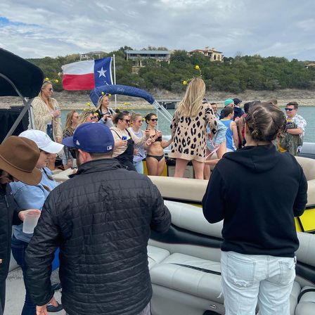 Pontoon boat party on a Texas lake — friends in swimsuits and casual jackets dancing and laughing under an overcast sky with a large Texas flag and shoreline homes in the background.