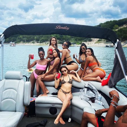 Seven women in bikinis posing with drinks on a pontoon boat during a sunny summer lake day with a wooded shoreline — lively boat party scene.