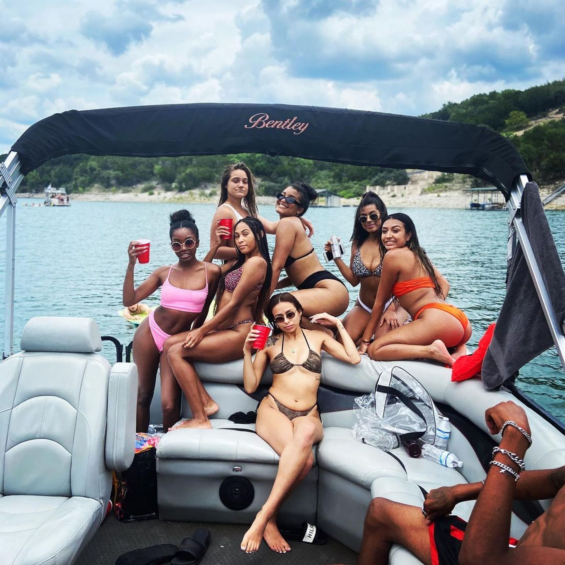 Seven women in bikinis posing with drinks on a pontoon boat during a sunny summer lake day with a wooded shoreline — lively boat party scene.