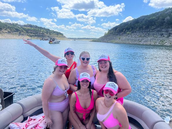 Six women in pink swimsuits and matching caps smiling on a pontoon boat at a cliff-lined lake under a sunny blue sky