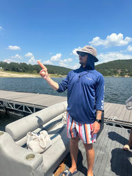 Man in a sun hat and sunglasses on a pontoon boat at a sunny lake dock, wearing a blue sun shirt and colorful striped swim trunks, playfully pointing toward the water with rolling hills and a bright blue sky in the background.