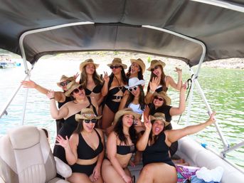Group of women in black swimsuits and straw cowboy hats laughing and posing on a covered pontoon boat over a green lake on a sunny summer day.
