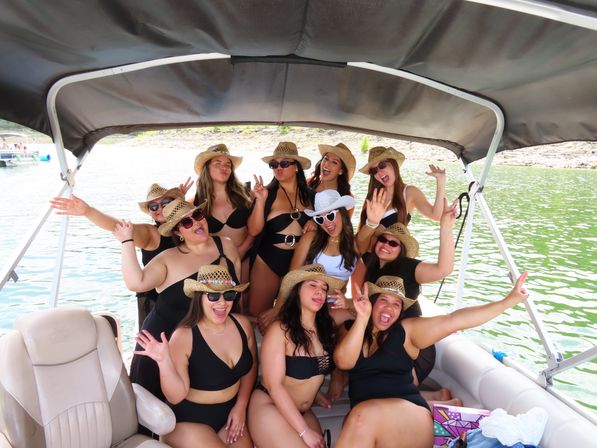 Group of women in black swimsuits and straw cowboy hats laughing and posing on a covered pontoon boat over a green lake on a sunny summer day.