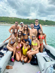 Group of friends in colorful swimsuits smiling and posing on a pontoon boat during a lake-day boat party, with green wooded shoreline and cloudy sky in the background.