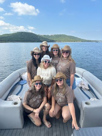 Seven friends wearing cowboy hats and matching Austin shirts smiling on a pontoon boat on a sunny lake with tree-covered hills in the background.